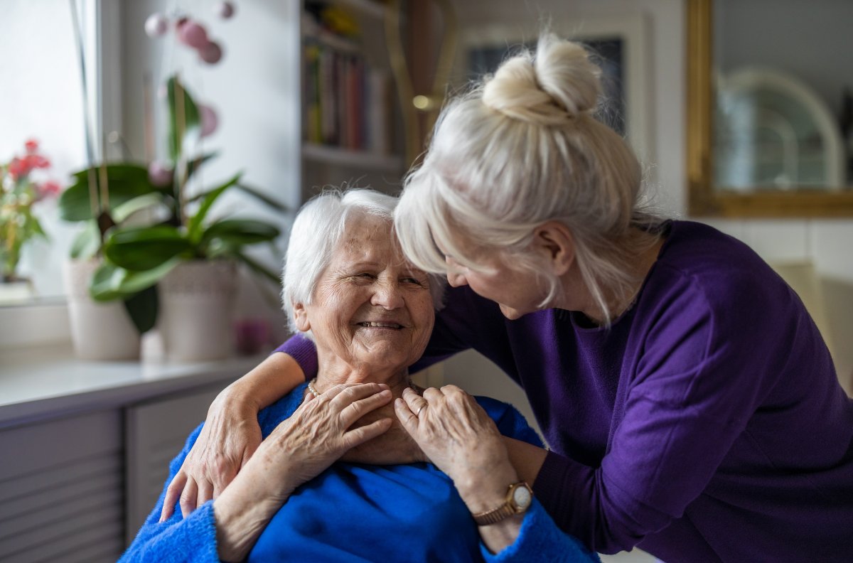 An older woman smiling as a family member leans in to embrace her in a sunlit living room